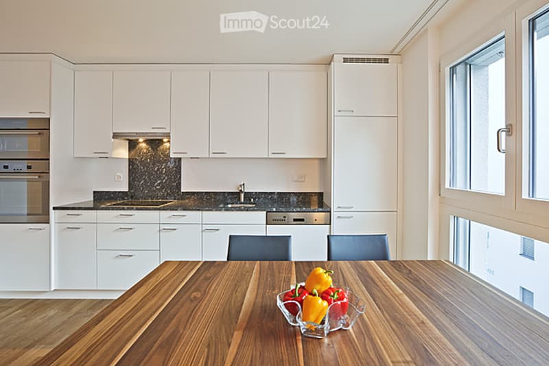 Modern kitchen with white cabinets, granite countertops, and stainless steel appliances. A wooden dining table with chairs is placed in the foreground, with a bowl of colorful bell peppers on it.
