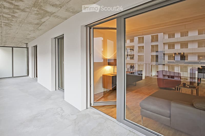 Modern, minimalist balcony with large glass doors opening to a furnished living space. The living area features a grey sofa, a wooden side table, a floor lamp, and a red armchair.