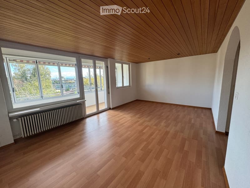 Empty room with wooden flooring, white walls, and large windows offering a view of trees and buildings outside. The room features a radiator under one window and a sliding glass door leading to another room.