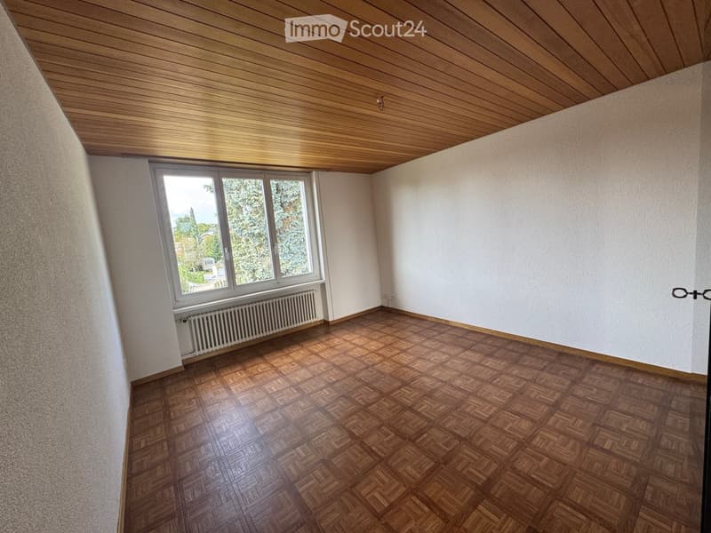 Empty room with wooden herringbone flooring, white walls, and a large window with a radiator below it. The ceiling has a wooden panel design.