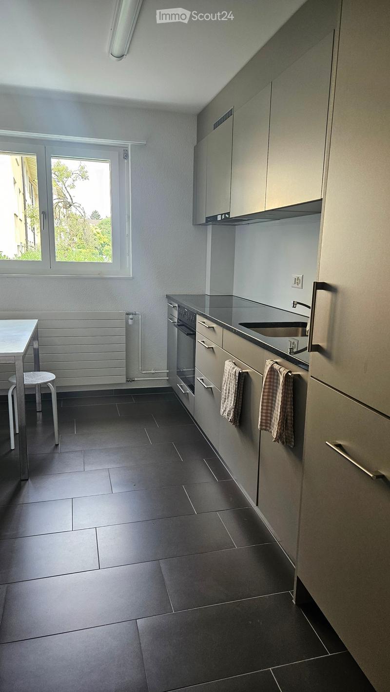 Modern kitchen with grey cabinets, dark tiled floor, stainless steel appliances, and a small table with a stool.