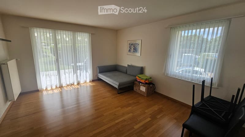 A modern living room with wooden flooring, large windows with sheer curtains, a grey sofa, a small table with folded blankets, and a stack of chairs.