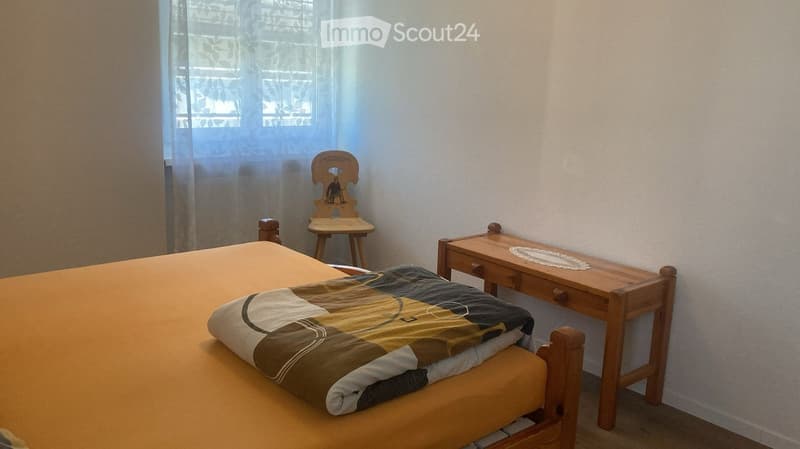 A small bedroom with a single bed, a wooden desk, and a wooden chair. The bed has a colorful quilt, and the desk has a doily on top.