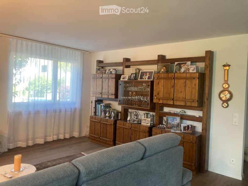 Living room with a grey sofa, coffee table, wooden bookshelf, and various decorative items including picture frames and glassware.