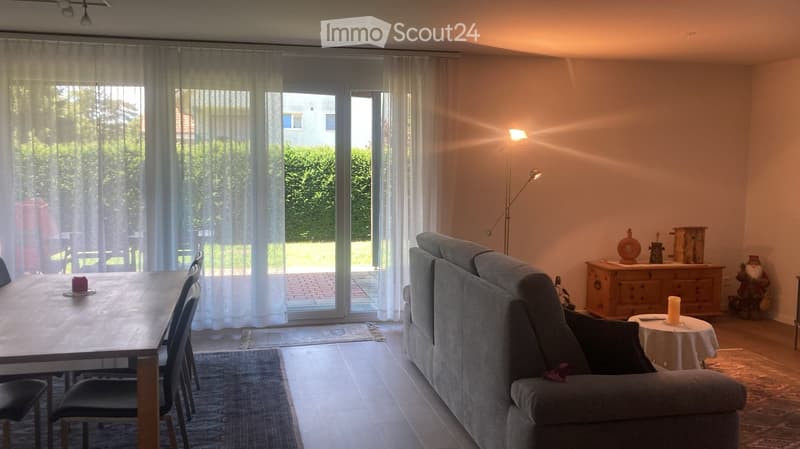 Bright living room with large sliding glass doors leading to a patio, a grey sofa, a wooden dining table with chairs, a floor lamp, and decorative items on a side table and cabinet.