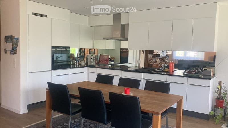 Modern kitchen with white cabinets, a wooden dining table with four chairs, a black countertop, and various appliances including an oven, coffee maker, and sink.