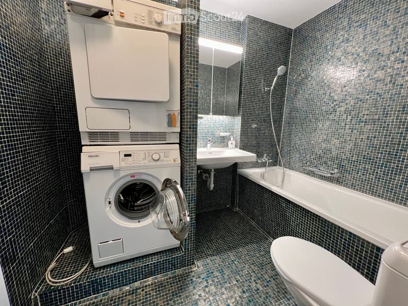 Bathroom with blue tiled walls and floor, featuring a toilet, bathtub, sink, and stacked washing machine and dryer unit.