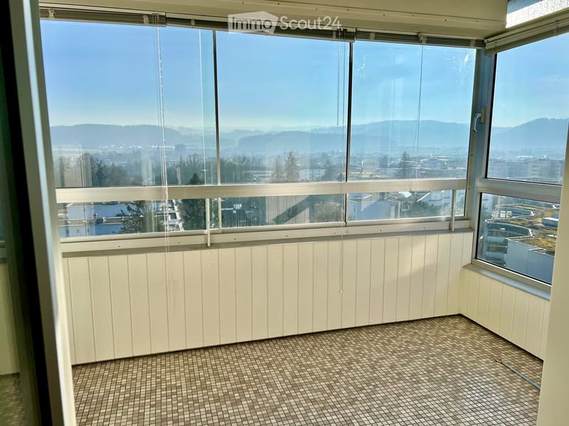 Empty room with large windows offering a view of the city and surrounding landscape, featuring tiled flooring and white paneled walls.