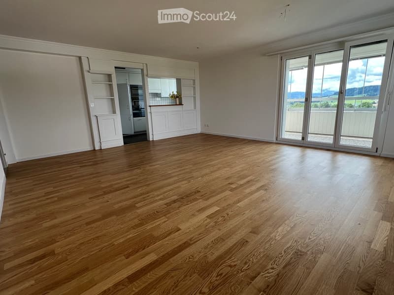 Empty room with wooden flooring, large sliding glass doors leading to a balcony, and a small kitchen area visible in the background.