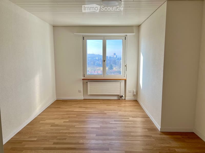 Empty room with wooden flooring, white walls, and a large window offering a city view. The room includes a radiator and a ceiling light fixture.