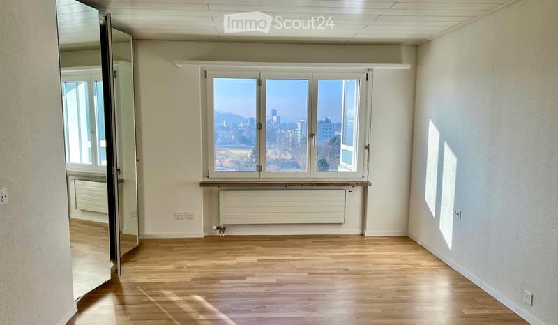 Empty room with wooden flooring, large windows offering a city view, and a radiator under the windows.