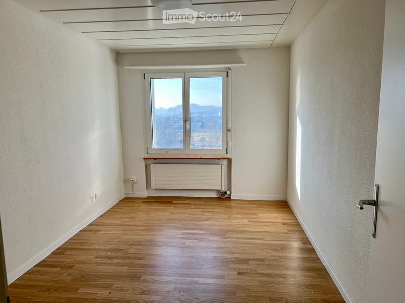 Empty hallway with wooden flooring, white walls, and a large window offering a view of the outside. A radiator is mounted below the window, and the ceiling features recessed lighting.