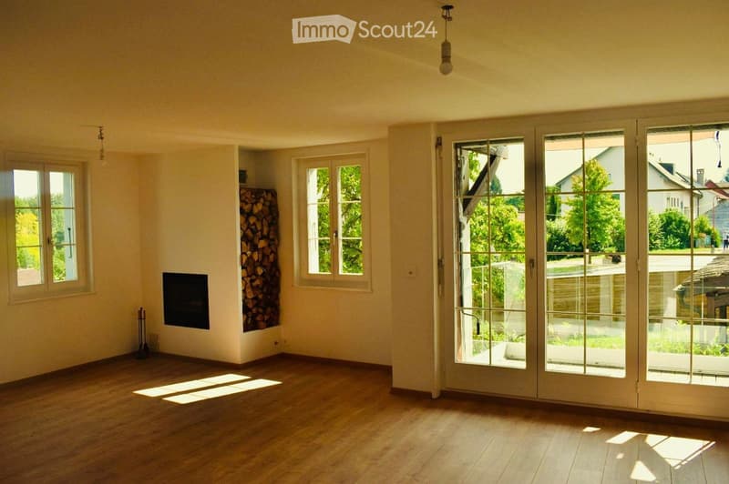 Empty living room with wooden flooring, large windows, and a fireplace. Natural light streams in, and there is a stack of firewood in one corner.