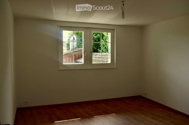 Empty room with wooden flooring and large windows offering a view of a tiled roof and greenery outside.