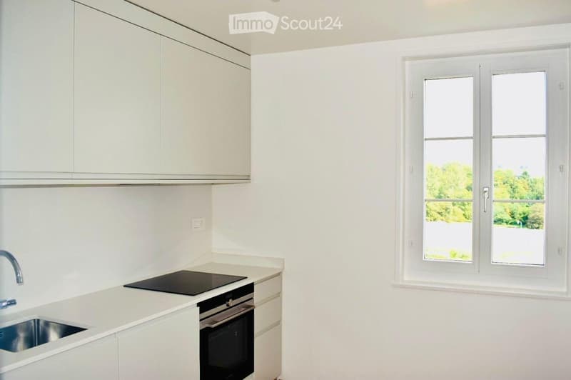 Modern kitchen with white cabinets, a sink, a faucet, and an oven. Large window with a view of greenery.