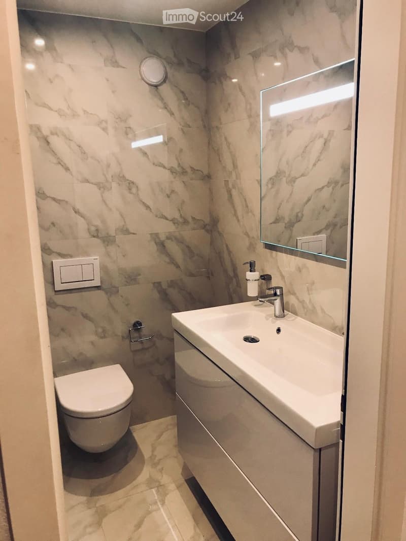 Modern bathroom with marble walls and floor, featuring a toilet, sink with modern faucet, and a large mirror with integrated lighting.