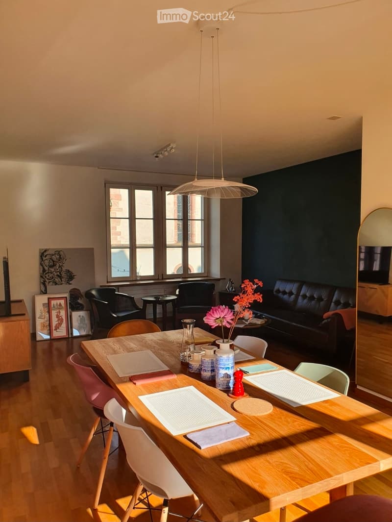 A modern, well-lit living and dining area with a wooden dining table, colorful chairs, a black leather sofa, and decorative items including a flower vase and picture frames.