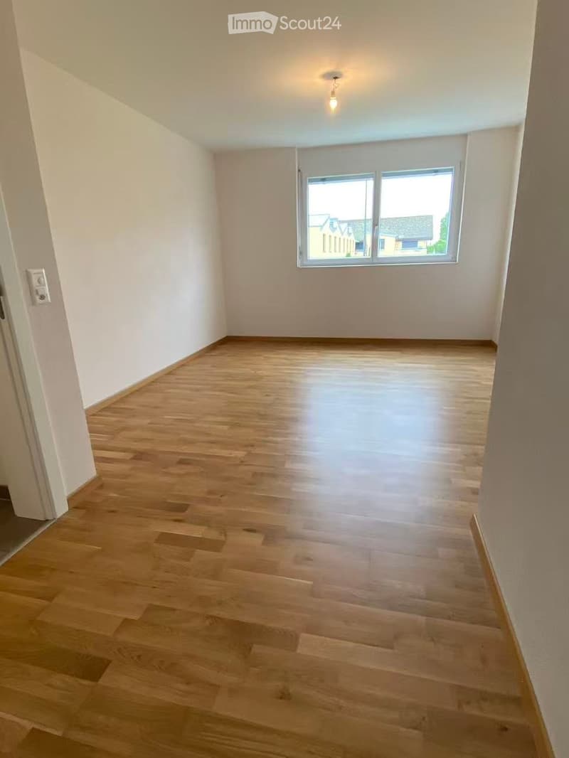 Empty room with wooden flooring, white walls, and a large window letting in natural light.