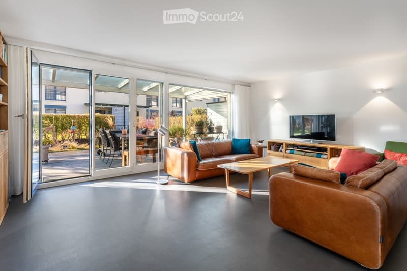 Modern living room with large glass sliding doors leading to a patio, leather sofas, a wooden coffee table, a television, and a floor lamp.