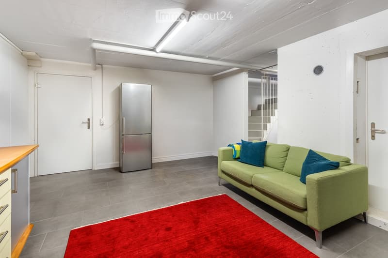 Modern living room with a green sofa, red rug, stainless steel refrigerator, and a staircase leading to another level.