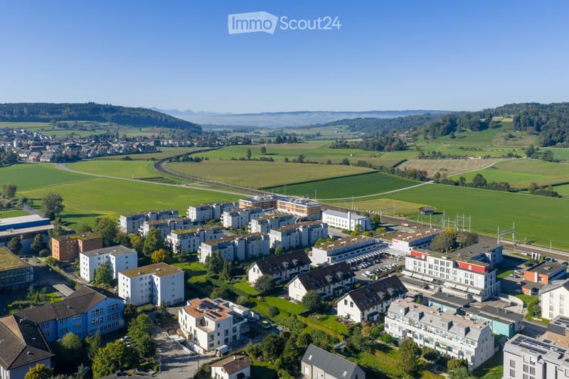 Aerial view of a residential area with multiple apartment buildings, parking lots, and surrounding green fields and forests.