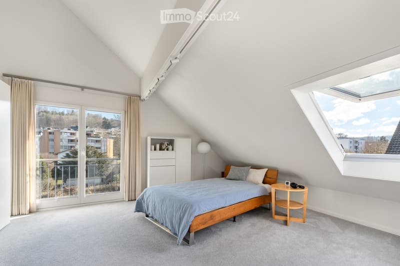 Bright attic bedroom with slanted ceilings, large windows, and a view of the city. Features a wooden bed, nightstand, cabinet, and floor lamp.