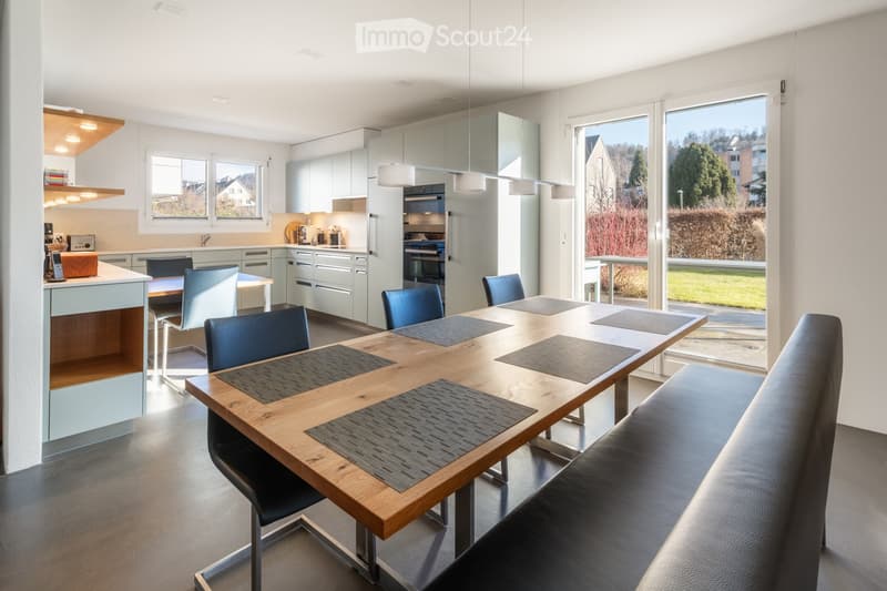 Modern kitchen with a light grey color scheme, featuring a central wooden dining table surrounded by blue chairs, a kitchen island with a countertop, and built-in cabinets with stainless steel appliances.