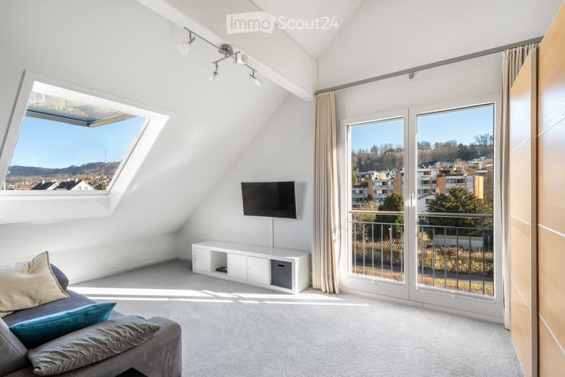 Bright, modern living room with a grey sofa, pillows, a flat-screen TV on a white cabinet, large windows with curtains, and a scenic view of the city and hills.