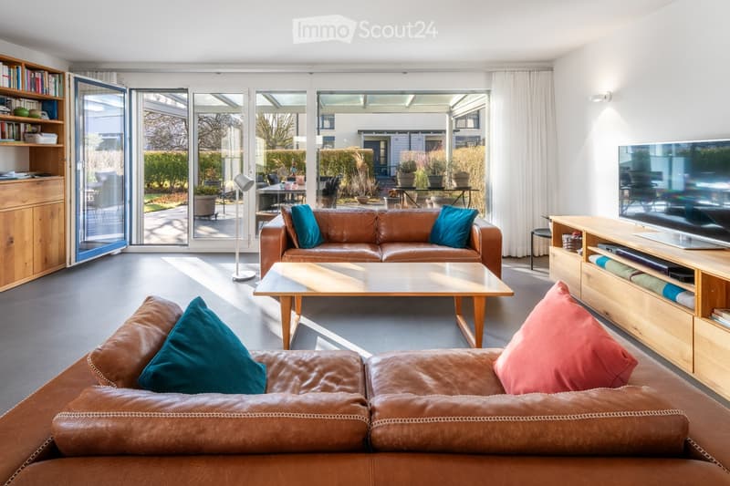 Bright living room with large glass doors leading to a patio, leather sofas, a coffee table, a TV stand, and a bookshelf.