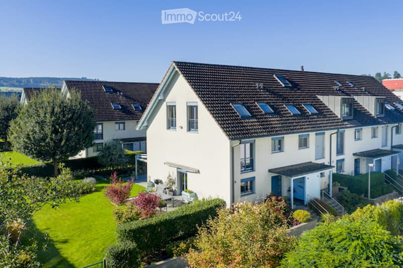 A modern two-story house with a dark tiled roof, white exterior walls, multiple windows, and a well-maintained garden with green lawns and shrubs.