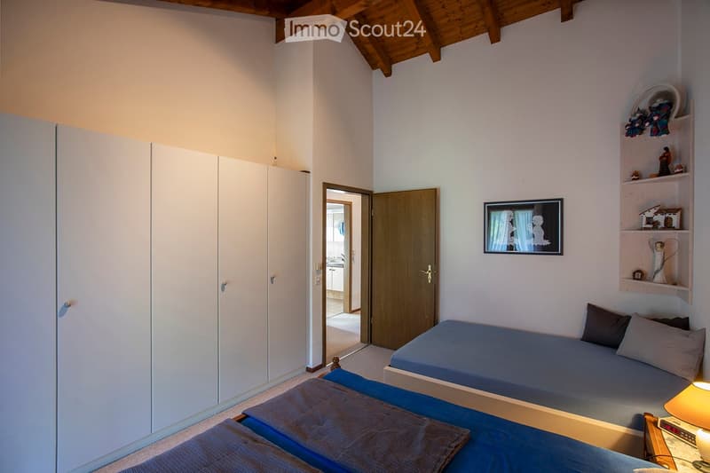 Two single beds, white walls, wooden door, built-in wardrobe, small wooden shelf with items, small framed artwork, nightstand with lamp