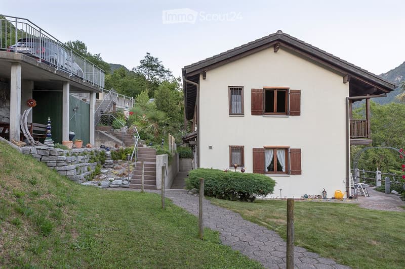 Two story house, raised ground floor, white walls, wooden windows, balcony, stairs leading to the entrance, pathway through the garden