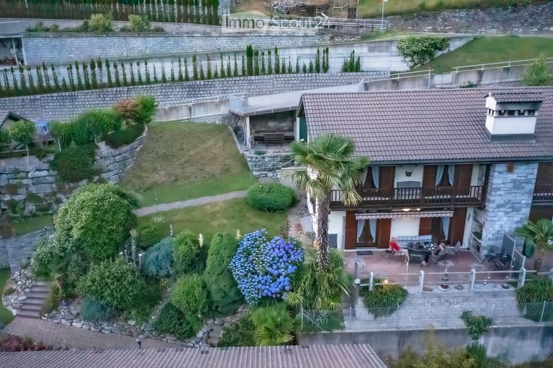 A two-story house with a tiled roof, surrounded by a well-maintained garden with various plants, trees, and a small patio area with outdoor furniture.