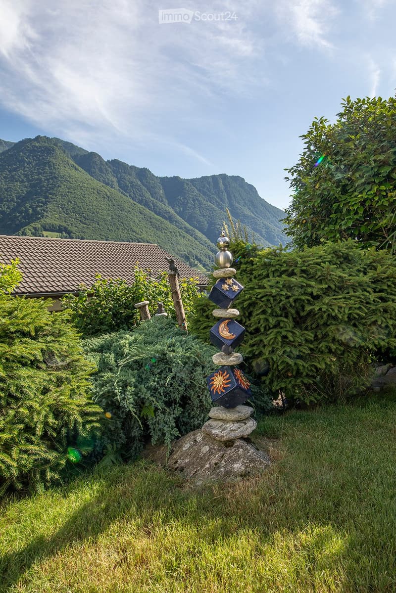 A garden with green lawn, various shrubs and trees, and a decorative stone pyramid with a celestial theme in the center, with mountains in the background