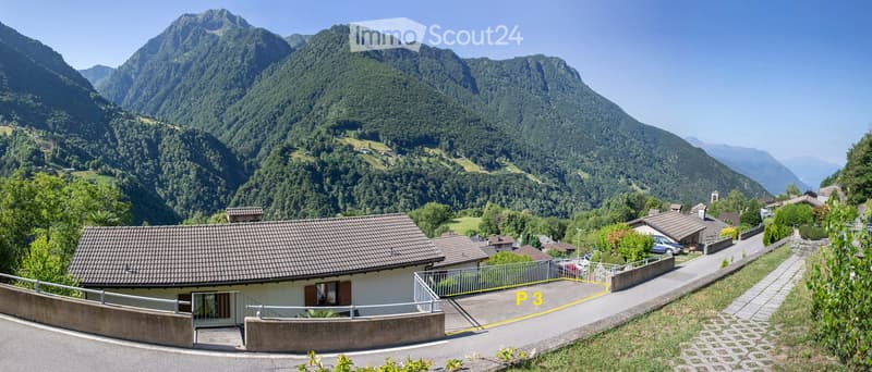 House with brown roof, surrounded by mountains, paved driveway, garden, and parking spot.