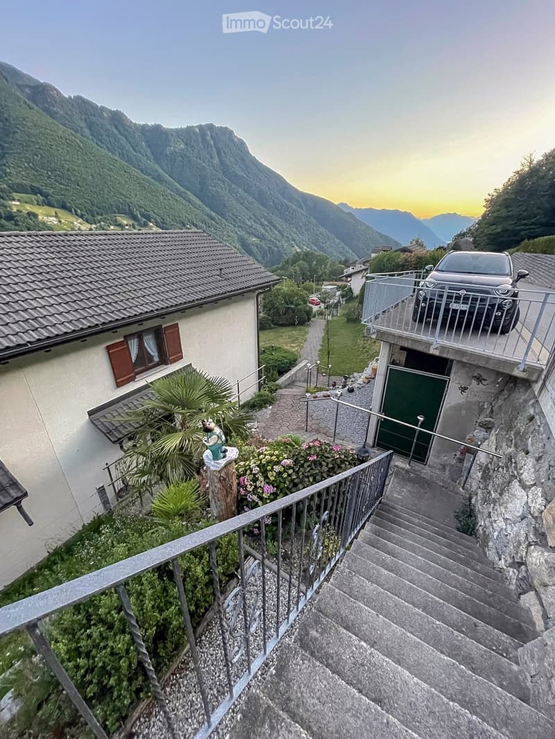 House on mountain side, steep stairs leading to the entrance, car parked on the balcony, house surrounded by green vegetation, mountain view