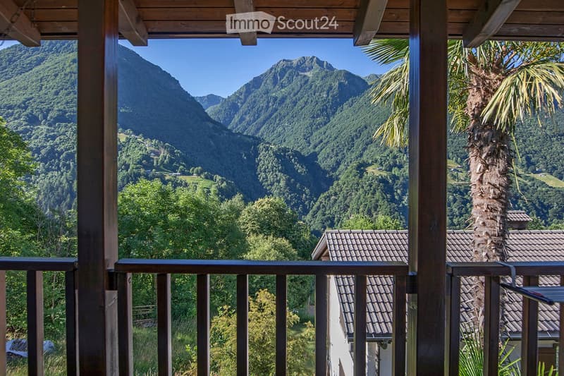 A balcony with a railing and wooden posts overlooking a scenic view of mountains and a house with a palm tree.