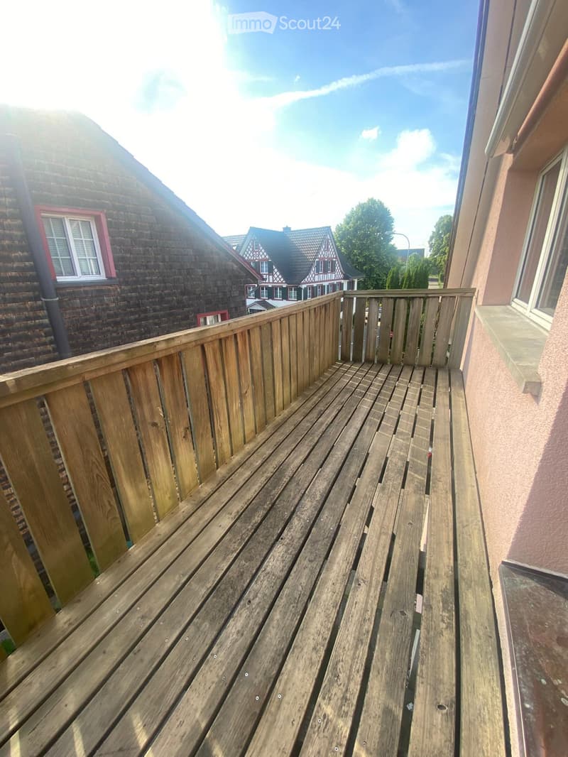 Wooden balcony with railing, overlooking a residential area with houses and trees.