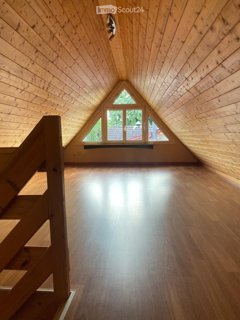 Bright, empty attic space with wooden walls, ceiling, and floor, featuring large triangular windows and a staircase leading up.