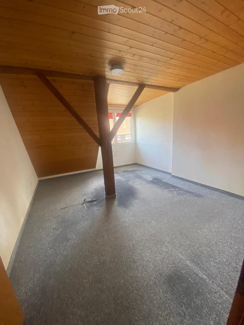 Empty room with wooden ceiling, exposed wooden beam, carpeted floor, and a small window with a view of another building.