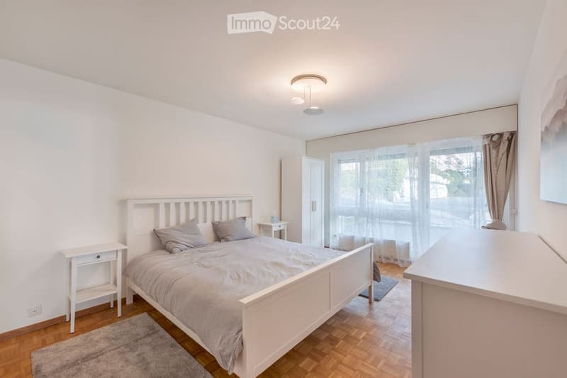 Bedroom. Bright bedroom with wooden flooring, white walls, and large windows. Features a white bed with grey bedding, two nightstands, a chair, and a mirror.