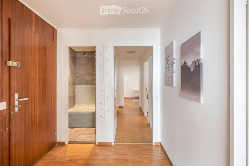Corridor / Hallway. Bright hallway with wooden flooring, white walls, and two open doors leading to other rooms. A small table and artwork are visible in the background.