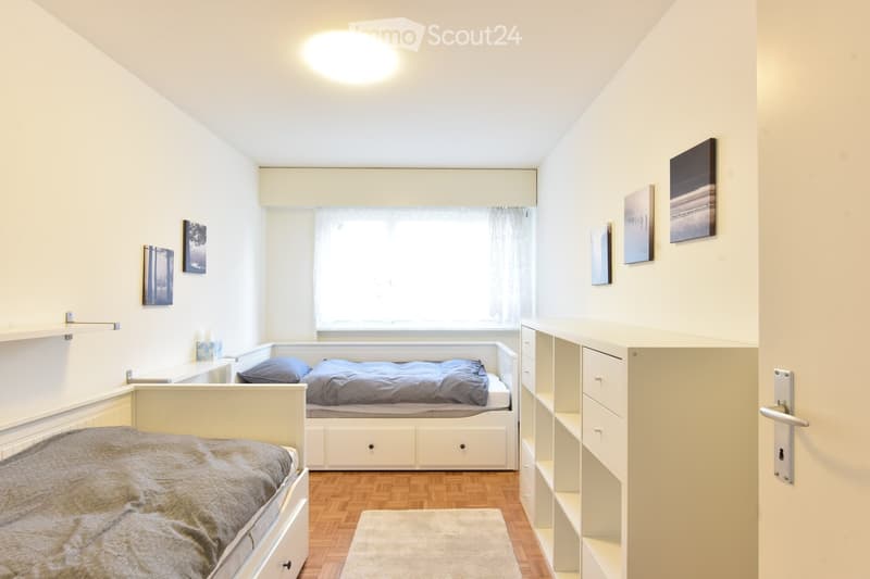 Bedroom. Cozy bedroom with two single beds, built-in wardrobe, shelves, and artwork on the walls. The room features wooden flooring and a large window with curtains.