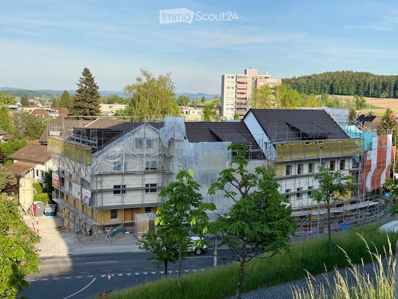 A multi-story building under construction with scaffolding and plastic sheeting covering parts of the facade. The building has a modern design with a dark roof and is surrounded by trees and other structures in the background.