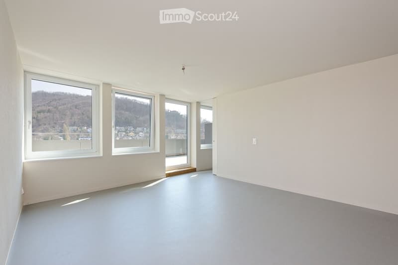 Empty room with light grey walls, grey floor, and large windows offering a scenic view of a town and hills.