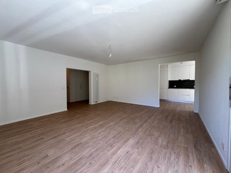 Empty room with wooden flooring, white walls, and a small adjacent kitchen area with white cabinets and a countertop.