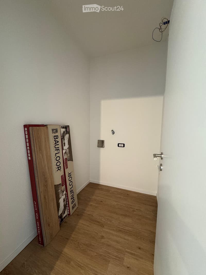 Empty hallway with wooden flooring, white walls, and a closed door at the end. There are some cardboard boxes and wooden planks leaning against the left wall.