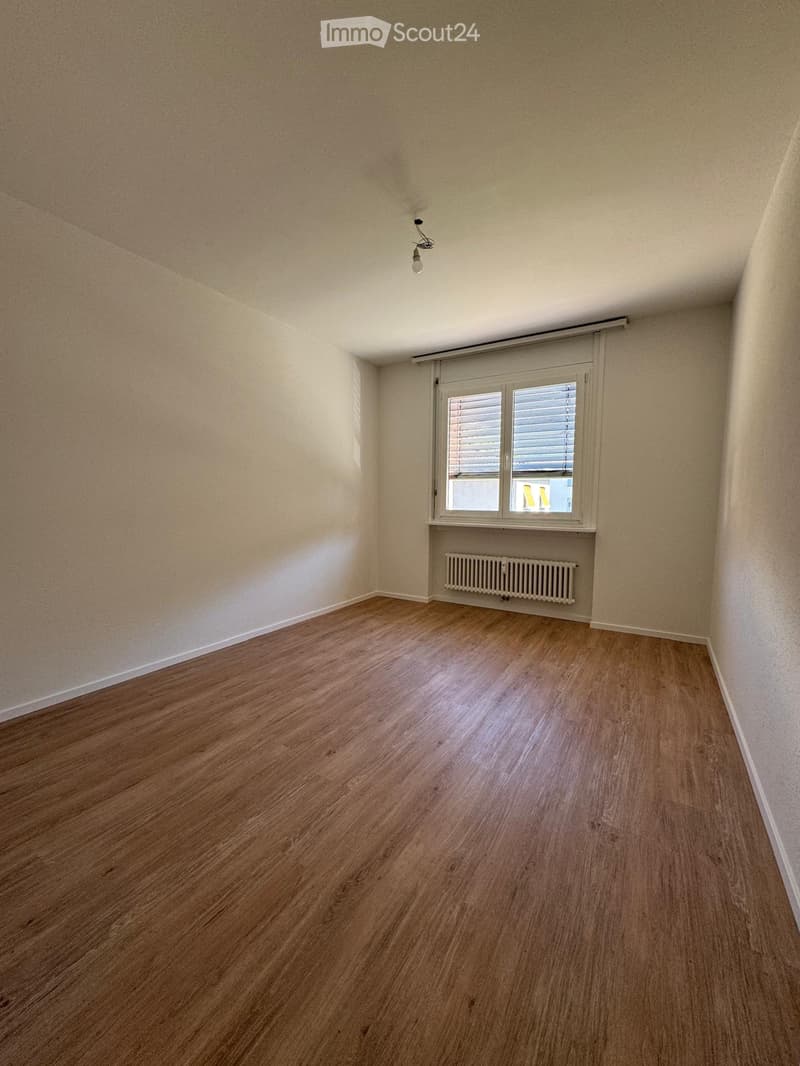 Empty room with wooden flooring, white walls, a window with blinds, and a radiator below the window.