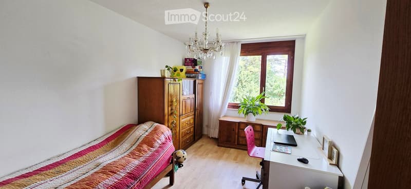 Cozy bedroom with a single bed, wooden wardrobe, desk with laptop, pink chair, and plants on the windowsill and shelf.