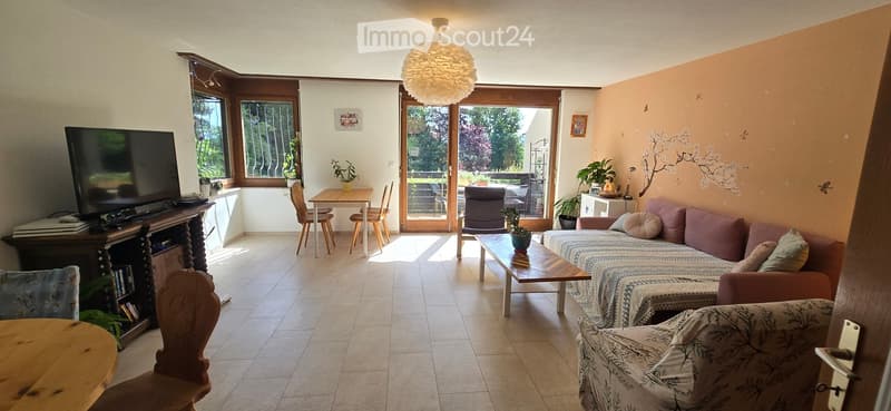 Bright living room with tiled floor, large windows, and a sliding glass door leading to a balcony. Features a patterned sofa, coffee table, dining table with chairs, and a television on a wooden stand.